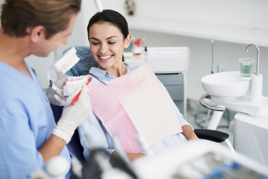 Consultation with doctor. Portrait of beautiful woman sitting in dental chair while stomatologist holding false teeth and toothbrush