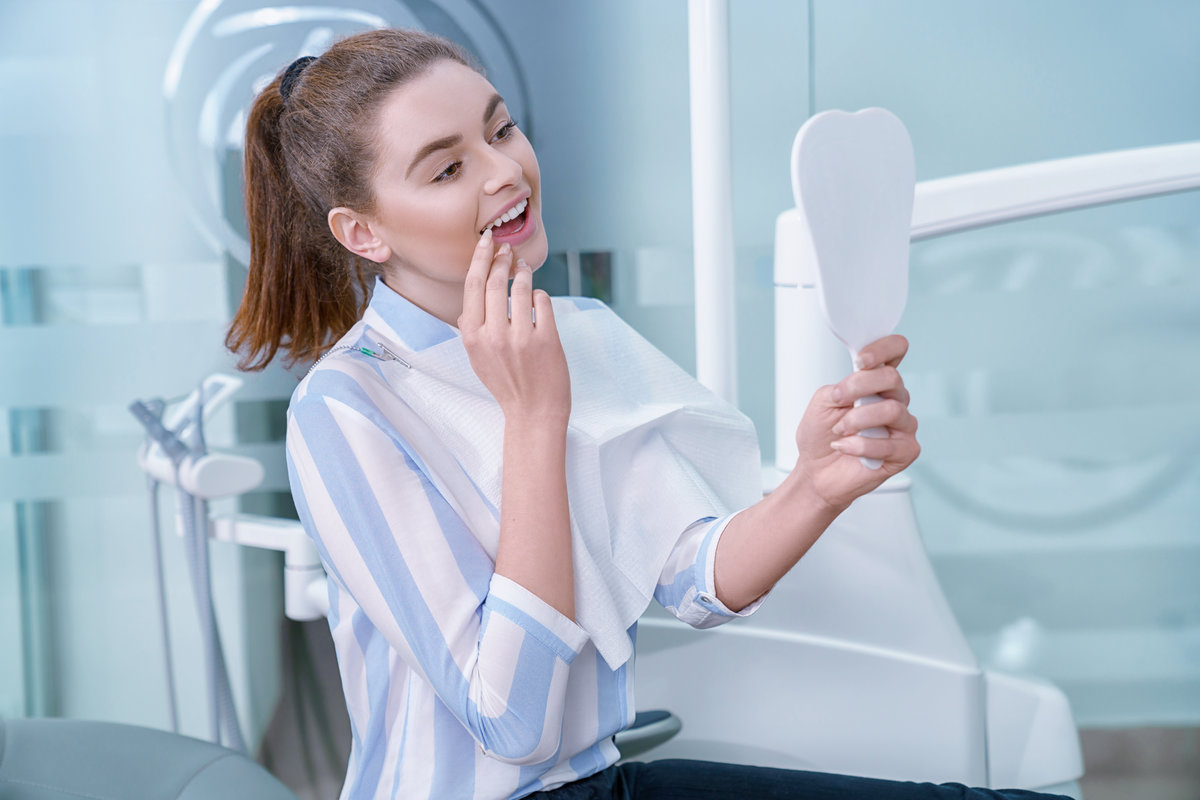 Happy woman satisfied with professional private clinic of dentistry. Charming and adorable woman looking at mirror with opened mouth, smiling. Client holding tooth shaped mirror.